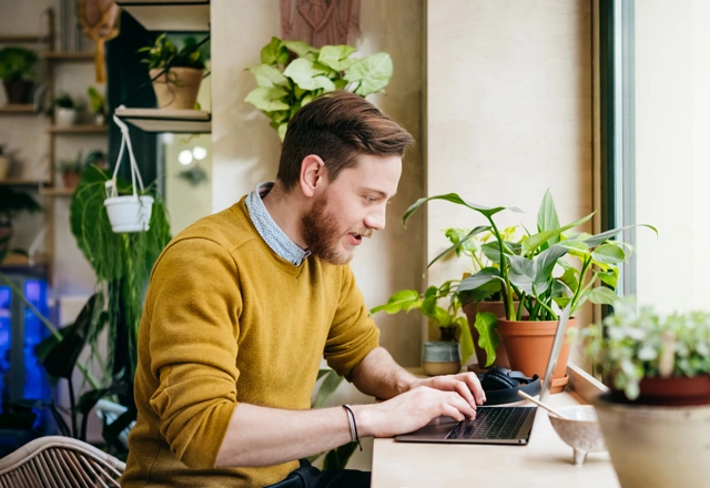 a man wearing a yellow jumper, sitting at a desk and working on laptop.