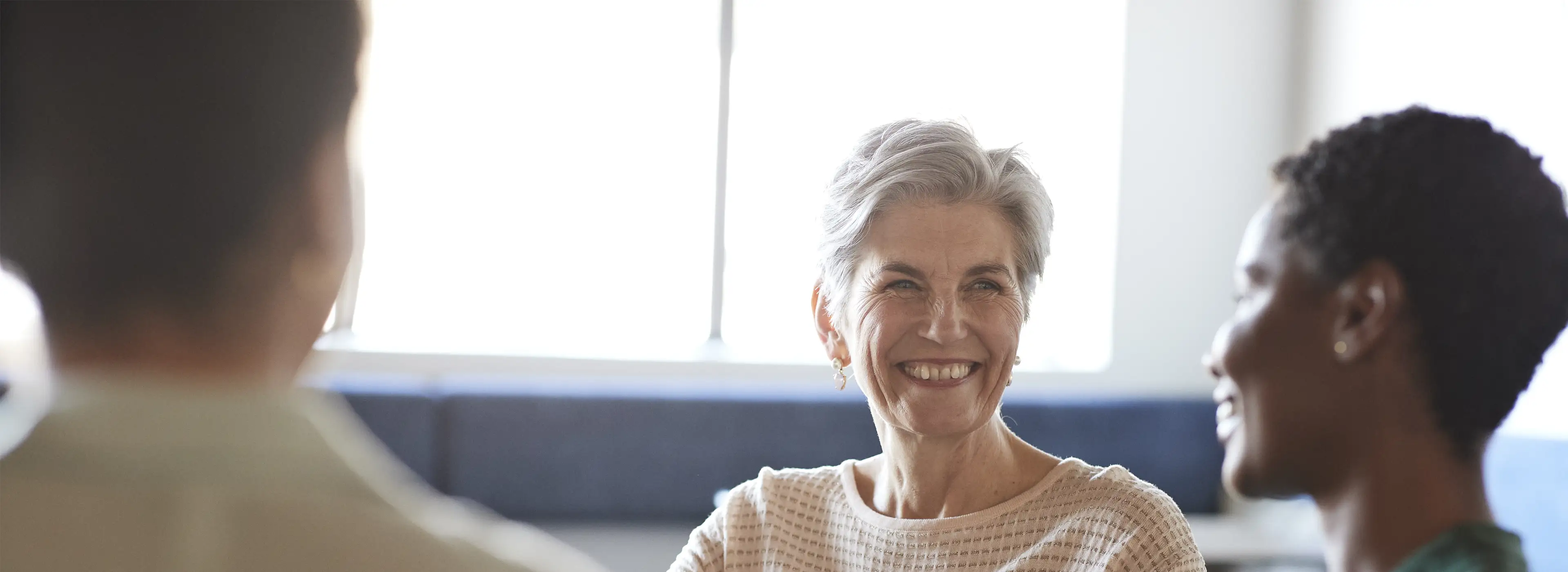 three people sitting a table talking. Woman facing the camera is wearing a white jumper and smiling. 